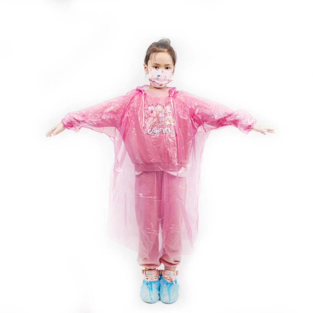 Child wearing pink raincoat and mask, arms outstretched.