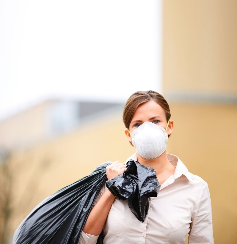 Person wearing mask, carrying garbage bag outdoors.